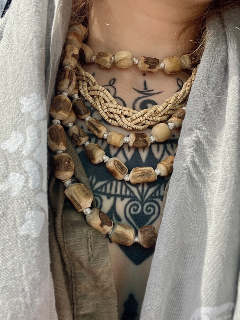 May include: A layered necklace made of multiple strands of wooden beads. The beads are a light brown colour and have a natural, rustic look. The necklace is worn over a brown shirt and a grey scarf.