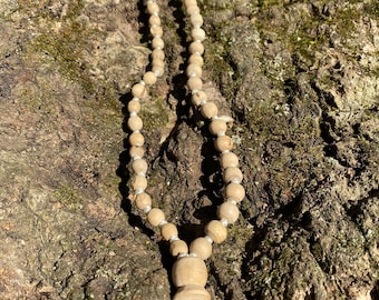 May include: A wooden mala necklace with a tassel, made of light brown beads, resting on a tree bark background.