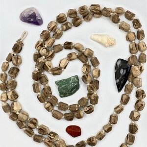 May include: A long strand of wooden beads in a light brown colour with a natural wood grain pattern. The beads are strung together with white string. There are several small stones and crystals scattered around the beads, including a purple amethyst, a green stone, a red stone, a white shell, and a black and white stone.
