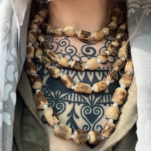 May include: A layered necklace made of natural wood beads. The beads are a light brown colour and have a rough, organic texture. The necklace is worn by a person with a black and white tattoo on their chest.