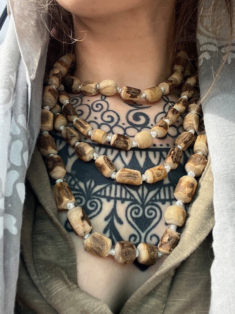 May include: A layered necklace made of natural wood beads. The beads are a light brown colour and have a rough, organic texture. The necklace is worn by a person with a black and white tattoo on their chest.