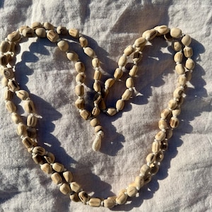 May include: A necklace made of light brown, oblong beads strung on white cord. The beads are arranged in a double strand, forming a heart shape. The necklace is displayed on a light-colored fabric.