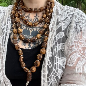 May include: A long, layered necklace made of brown wooden beads. The necklace is worn by a person with tattoos on their chest.