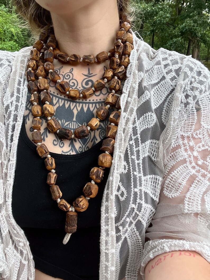 May include: A long, layered necklace made of brown wooden beads. The necklace is worn by a person with tattoos on their chest.