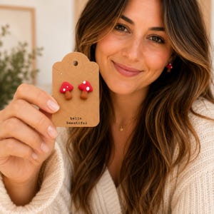 May include: A pair of red mushroom earrings with white spots on a brown tag that says "hello beautiful". The earrings are displayed on a brown tag. The earrings are red with white spots and brown stems.