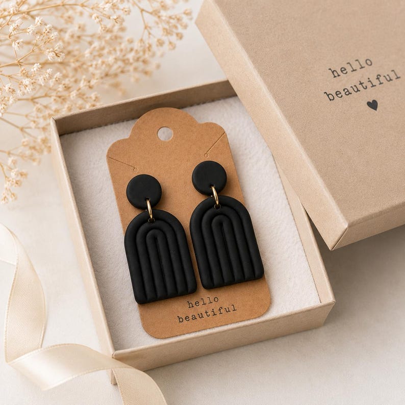 K&ouml;nnte beinhalten: Ein Paar mattschwarze, bogenf&ouml;rmige Ohrringe mit gerippter Textur, befestigt an einer braunen Pappkarte mit dem Aufdruck hello beautiful. Die Ohrringe befinden sich in einer passenden braunen Geschenkbox mit demselben Text.