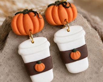 May include: A pair of dangle earrings featuring an orange pumpkin top connected to a white coffee cup with a brown sleeve. Each sleeve is decorated with a small orange pumpkin accent. The earrings are attached with gold-toned metal jump rings.