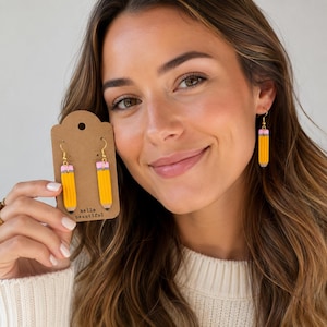 May include: Dangle earrings shaped like yellow pencils with pink erasers and grey tips. The earrings feature gold-toned metal hooks. One pair is attached to a brown cardboard display card printed with the text hello beautiful at the bottom.