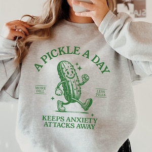 May include: A gray sweatshirt with a green graphic of a walking pickle. The graphic says "A Pickle A Day" with "More Dill" and "Less Pills" on either side of the pickle. The graphic also says "Keeps Anxiety Attacks Away".