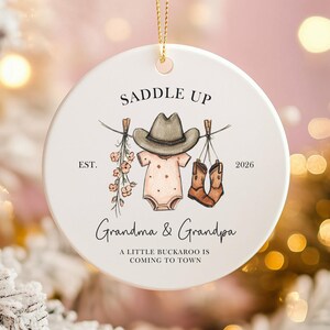 May include: A white ceramic Christmas ornament with a gold string. The ornament features a cowboy hat, baby clothes, and boots hanging on a clothesline. Text includes "SADDLE UP", "Grandma & Grandpa", and "A LITTLE BUCKAROO IS COMING TO TOWN".