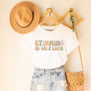May include: White t-shirt with the text "Stimming is Self Care" in colorful, rounded letters. The shirt is displayed on a hanger with a straw hat and a woven bag.