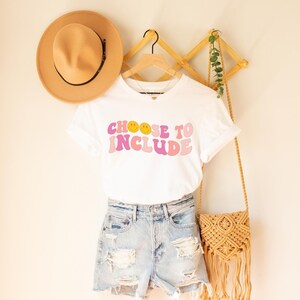 May include: White t-shirt with the text "Choose to Include" in pink and yellow letters with smiley faces. The shirt is hanging on a wooden rack with a straw hat and a woven bag.