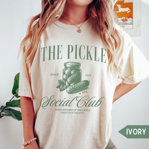 May include: A cream-colored t-shirt with a green graphic of a jar of pickles and the text "THE PICKLE Social Club SINCE 1923 PROUD MEMBER OF THE PICKLE ADDICTION SOCIETY."