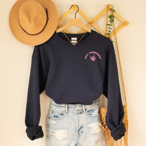 May include: A navy blue sweatshirt with a pink embroidered design that says "X-Ray Technologist" with a hand gesture. The sweatshirt is hanging on a wooden hanger and is paired with light blue denim shorts.