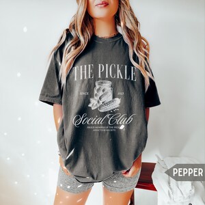 May include: A dark gray t-shirt with a white graphic design that says "The Pickle Social Club" with a jar of pickles and the text "Since 1923" and "Proud Member of the Pickle Addiction Society".