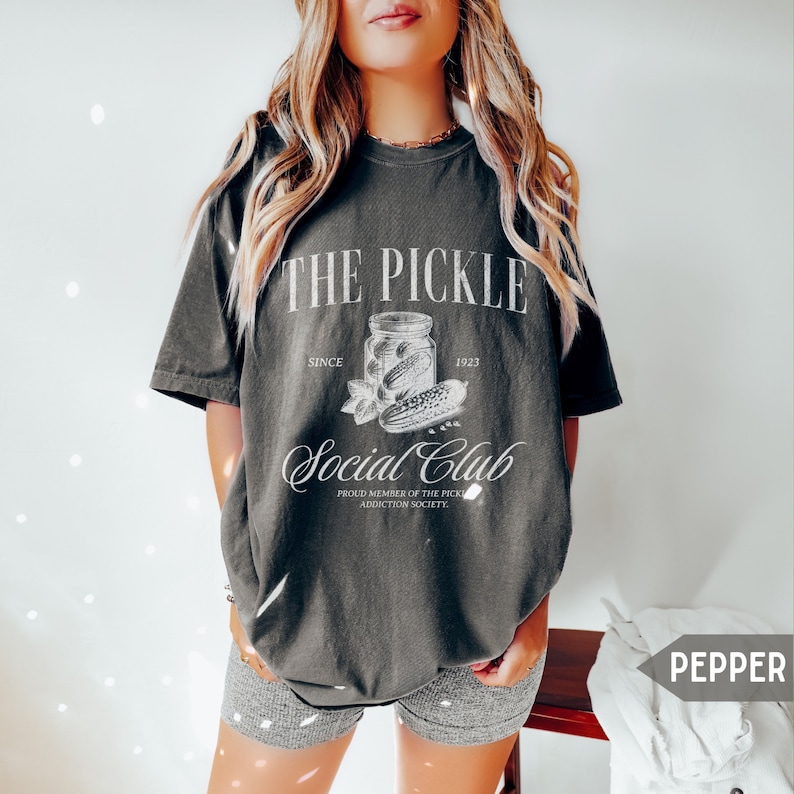 May include: A dark gray t-shirt with a white graphic design that says "The Pickle Social Club" with a jar of pickles and the text "Since 1923" and "Proud Member of the Pickle Addiction Society".