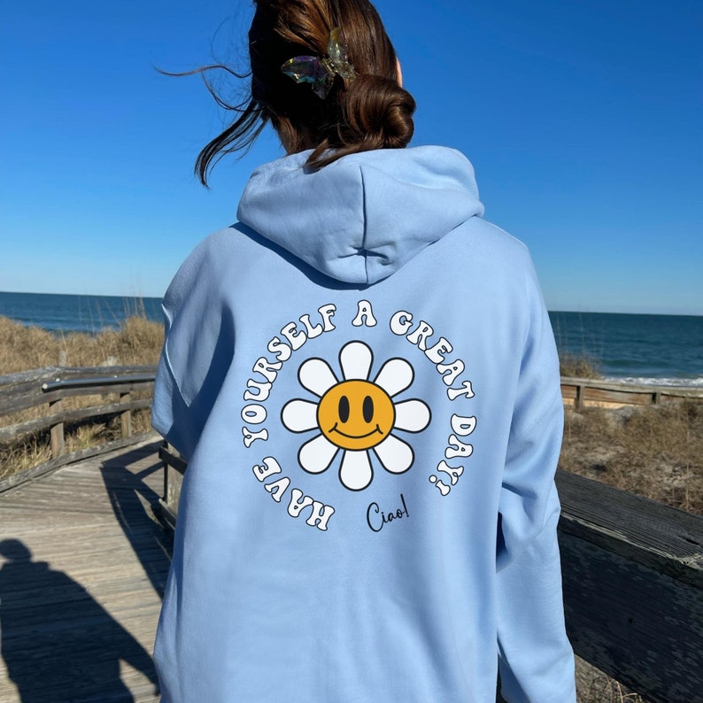 May include: A light blue hoodie with a white daisy graphic on the back. The daisy has a yellow smiley face in the center and the text "Have yourself a great day! Ciao!" around it.