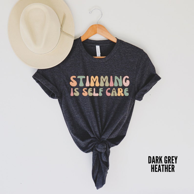 May include: Dark grey heather t-shirt with the text "Stimming is Self Care" in colourful, rounded letters.