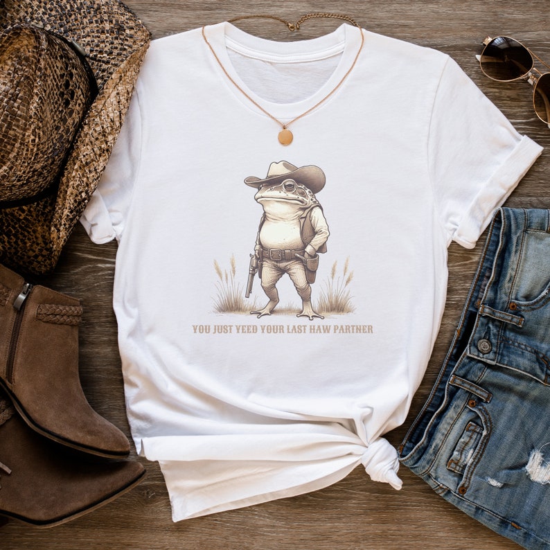 May include: White t-shirt featuring a cartoon image of a frog wearing a cowboy hat and holding a gun. The text "You just yeed your last haw partner" is printed below the image.