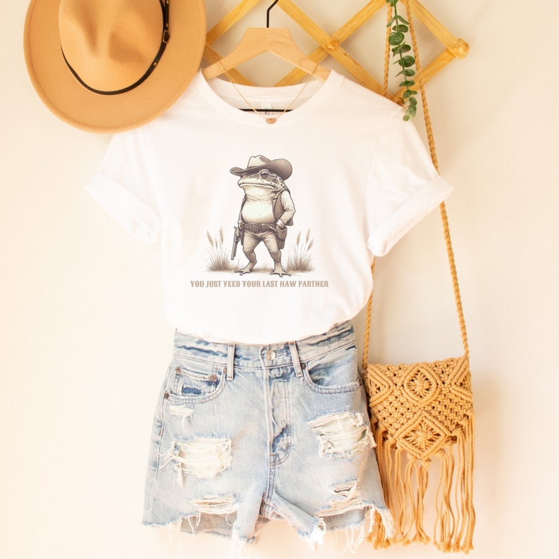 May include: White t-shirt featuring a cartoon illustration of a frog wearing a cowboy hat and holding a gun. The text "You just yeed your last haw partner" is printed below the image.