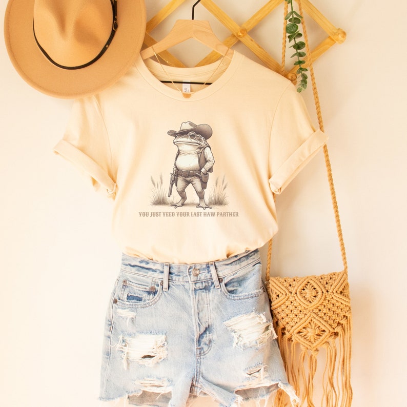 May include: A beige t-shirt featuring a graphic of a frog wearing a cowboy hat and holding a gun. The text "You just yeed your last haw partner" is printed below the frog.