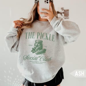 May include: Ash gray sweatshirt with the text "THE PICKLE Social Club" in green. The design includes a jar of pickles, a cucumber, and the text "Since 1923". The sweatshirt is a casual style.