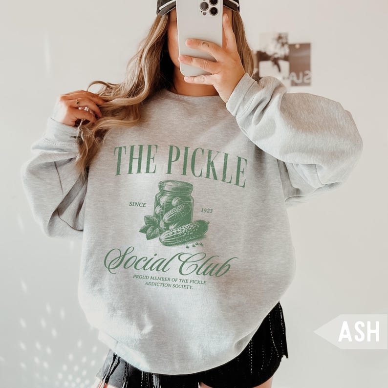 May include: Ash gray sweatshirt with the text "THE PICKLE Social Club" in green. The design includes a jar of pickles, a cucumber, and the text "Since 1923". The sweatshirt is a casual style.