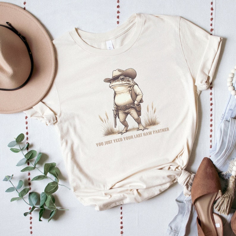 May include: A beige t-shirt featuring a cartoon illustration of a frog wearing a cowboy hat and holding a gun. The text "You just yeed your last haw partner" is printed below the frog.