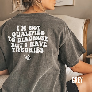 May include: A grey t-shirt with the text "I'm not qualified to diagnose but I have theories" printed on the back. The text is in a white, retro font with a skeleton hand giving the middle finger below it.