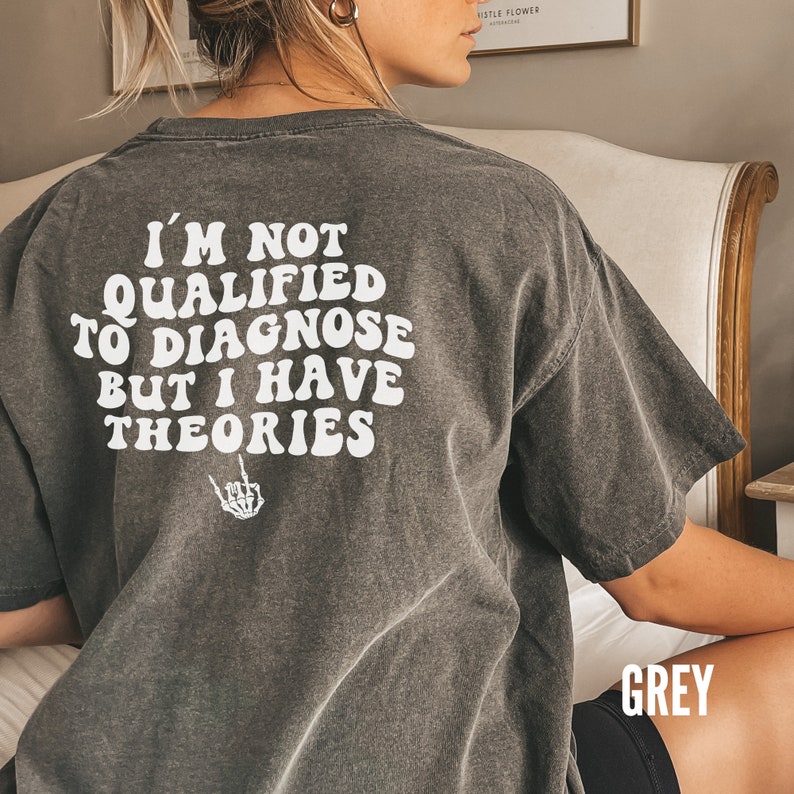 May include: A grey t-shirt with the text "I'm not qualified to diagnose but I have theories" printed on the back. The text is in a white, retro font with a skeleton hand giving the middle finger below it.