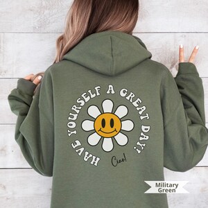 May include: A green hooded sweatshirt with a white daisy with a yellow smiley face in the center. The text "Have yourself a great day! Ciao!" is printed in a retro font around the daisy.