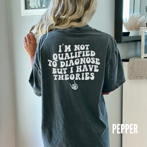 May include: A dark grey t-shirt with white text that reads "I'm not qualified to diagnose but I have theories" with a skeleton hand giving the middle finger.