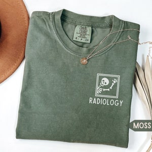 May include: A moss green t-shirt with a white graphic of a skeleton holding a bone and the text "RADIOLOGY".