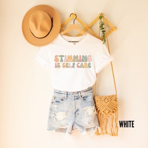 May include: White t-shirt with the text "Stimming is Self Care" in colourful, rounded letters. The shirt is hanging on a wooden rack with a straw hat and a woven bag.