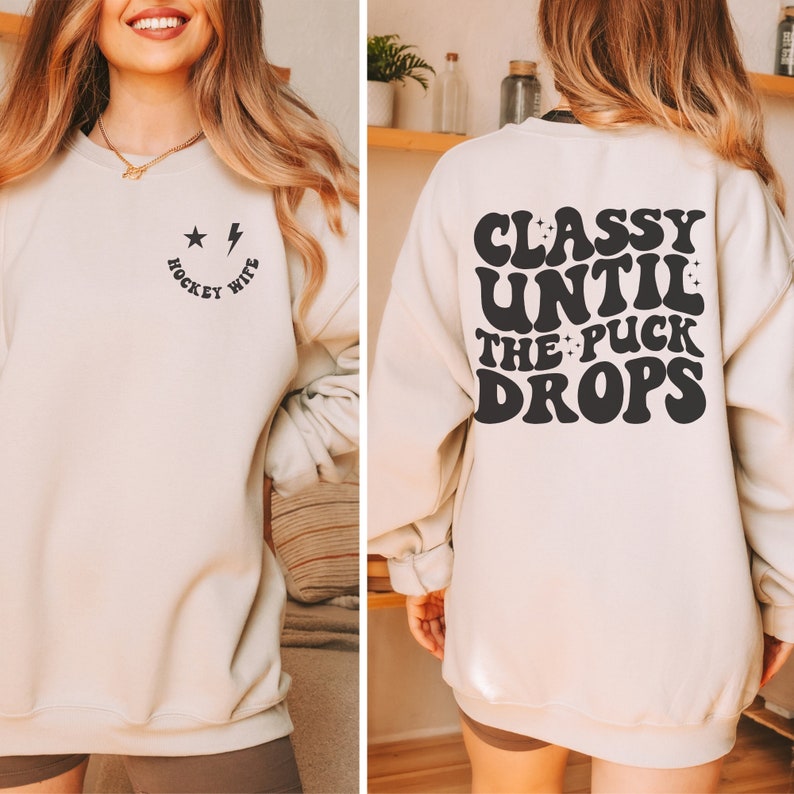 Pu&ograve; includere: Una felpa beige chiaro con una grafica nera sul davanti che dice "Hockey Wife" con una faccina sorridente, una stella e un fulmine. La parte posteriore della felpa ha una grafica nera che dice "Classy Until The Puck Drops" in un font retr&ograve;.