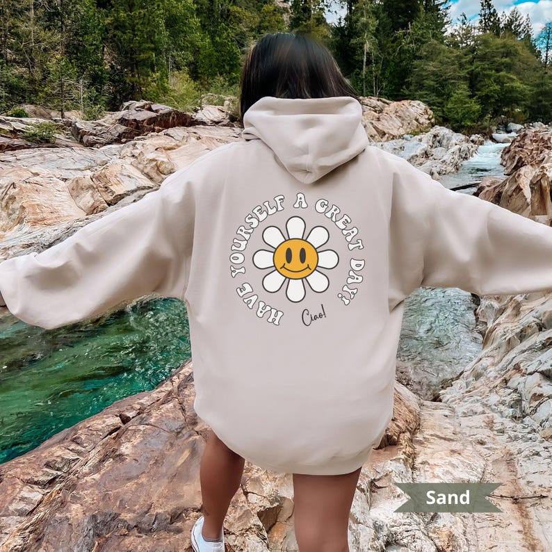 May include: A beige hoodie with a graphic of a yellow smiley face flower on the back. The text "Have yourself a great day! Ciao!" is printed around the flower.