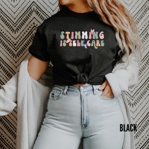 May include: A black t-shirt with the text "Stimming is self care" in rainbow colors with butterfly accents. The shirt is worn by a person with long brown hair and is tied at the waist.