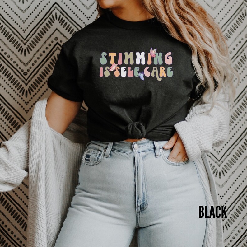 May include: A black t-shirt with the text "Stimming is self care" in rainbow colors with butterfly accents. The shirt is worn by a person with long brown hair and is tied at the waist.