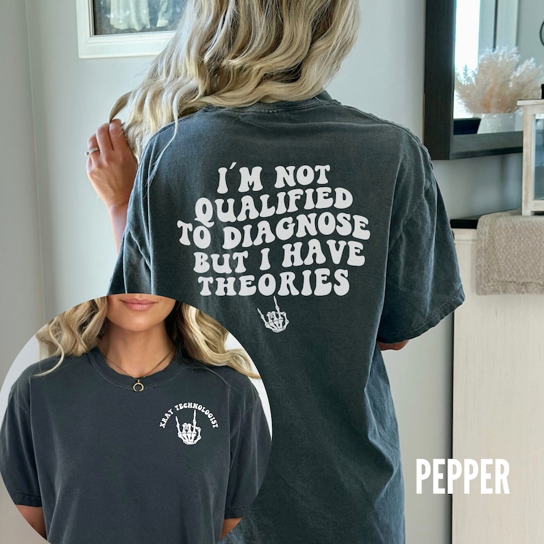 May include: A dark gray t-shirt with the text "I'm not qualified to diagnose but I have theories" in white. The text is in a retro font style. A skeleton hand with a middle finger is below the text. The t-shirt has a crew neck and short sleeves. The word "Pepper" is in white text on a black background in the lower right corner of the image.