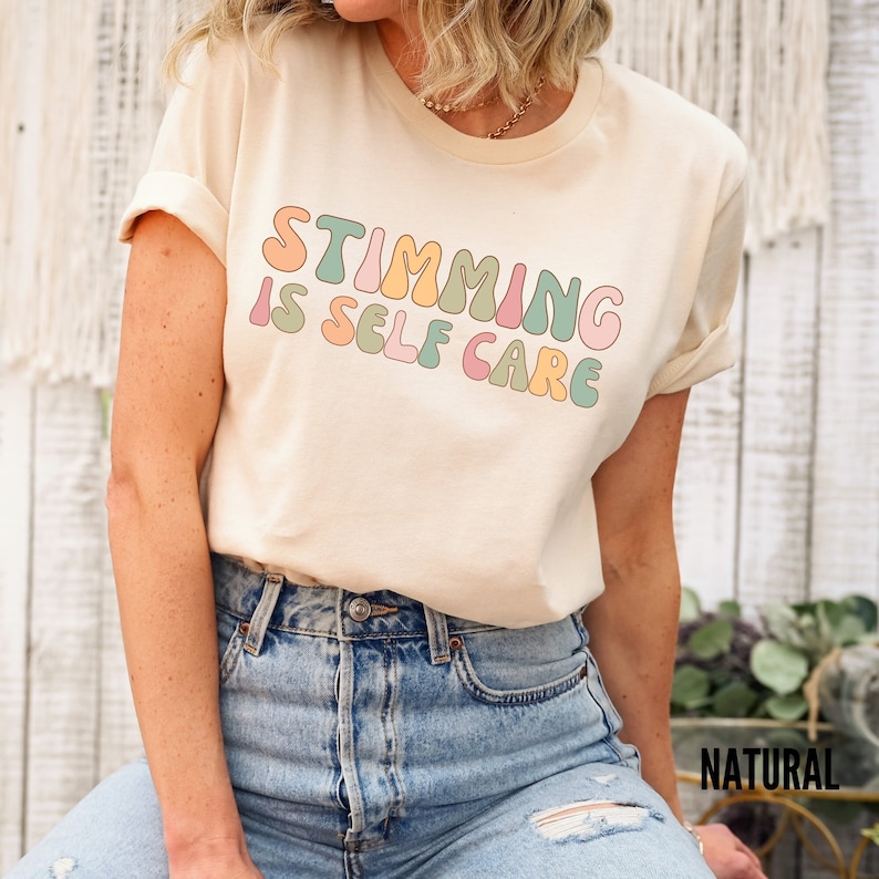 May include: A light beige t-shirt with the text "Stimming is Self Care" in colourful, rounded letters.