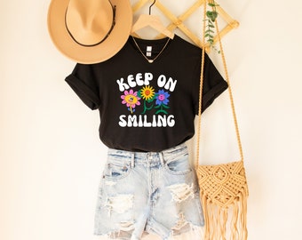 May include: Black t-shirt with a graphic of three smiling flowers and the text "Keep on Smiling". The shirt is hanging on a wooden rack with a straw hat and a woven bag.