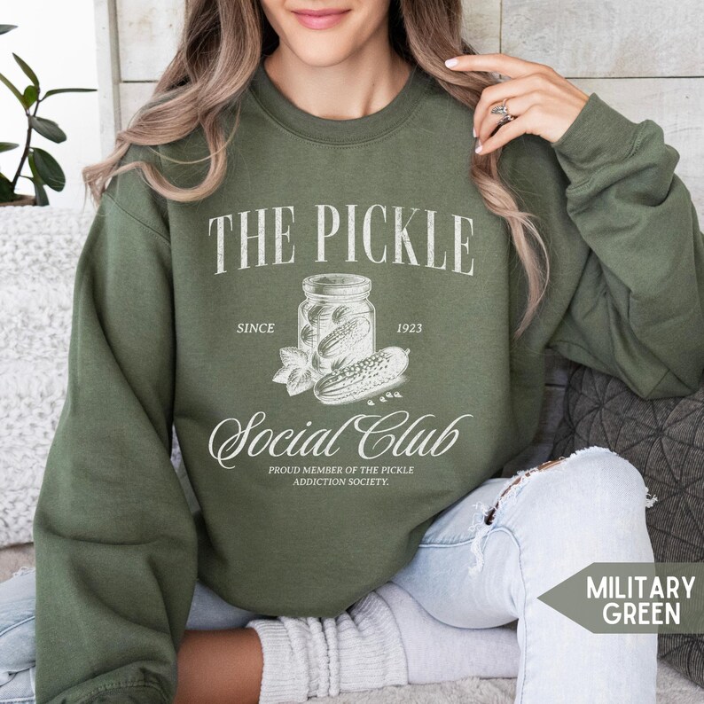 May include: Military green sweatshirt with the text "THE PICKLE Social Club" and a graphic of a pickle jar. The design includes the text "SINCE 1923" and "PROUD MEMBER OF THE PICKLE ADDICTION SOCIETY."