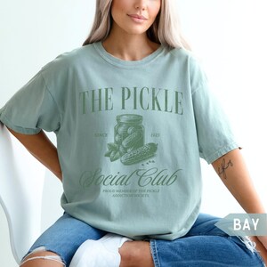 May include: A light blue t-shirt with a green graphic of a jar of pickles and the text "THE PICKLE Social Club SINCE 1923 PROUD MEMBER OF THE PICKLE ADDICTION SOCIETY."