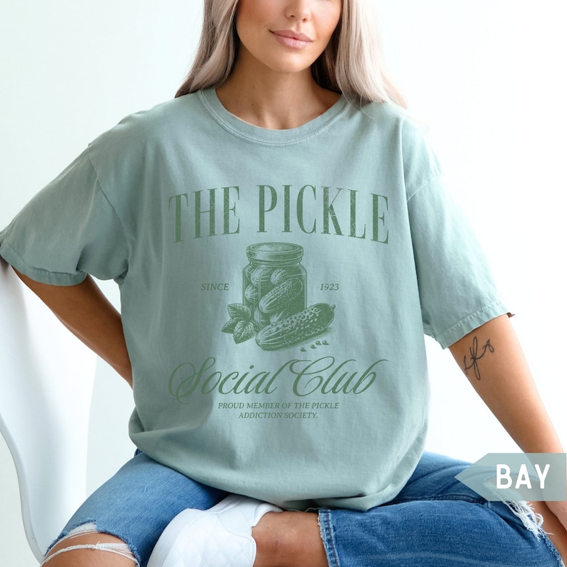 May include: A light blue t-shirt with a green graphic of a jar of pickles and the text "THE PICKLE Social Club SINCE 1923 PROUD MEMBER OF THE PICKLE ADDICTION SOCIETY."