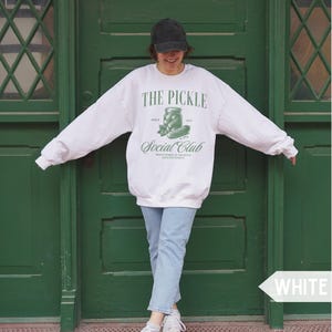 May include: White sweatshirt with the text "THE PICKLE Social Club" in green. The design includes a pickle jar and cucumber illustration. The person in the image is wearing a black cap and light blue jeans.