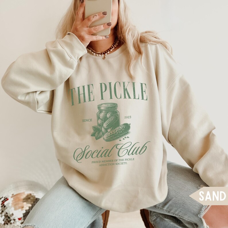 May include: Sand-colored sweatshirt with the text "THE PICKLE Social Club" in green. The design includes a jar of pickles and the text "SINCE 1923". The sweatshirt is a comfortable, casual style.
