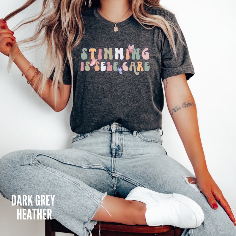 May include: A dark grey heather t-shirt with the text "Stimming is Self Care" in rainbow colors. The text is in a retro font style with butterflies surrounding it.