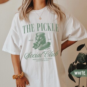 May include: White t-shirt with a green graphic of a jar of pickles and the text "THE PICKLE SINCE 1923 Social Club PROUD MEMBER OF THE PICKLE ADDICTION SOCIETY."