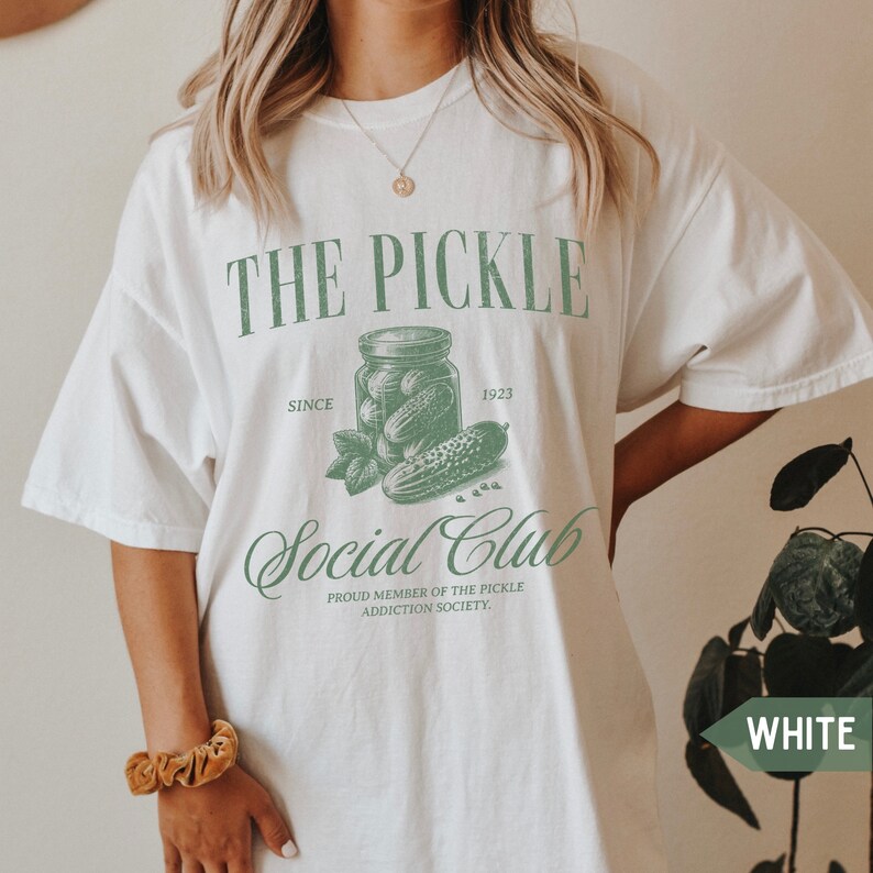 May include: White t-shirt with a green graphic of a jar of pickles and the text "THE PICKLE SINCE 1923 Social Club PROUD MEMBER OF THE PICKLE ADDICTION SOCIETY."