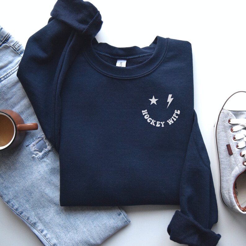 Pu&ograve; includere: Felpa blu navy con un grafico di una faccia sorridente bianca e il testo "Hockey Wife" sul davanti.
